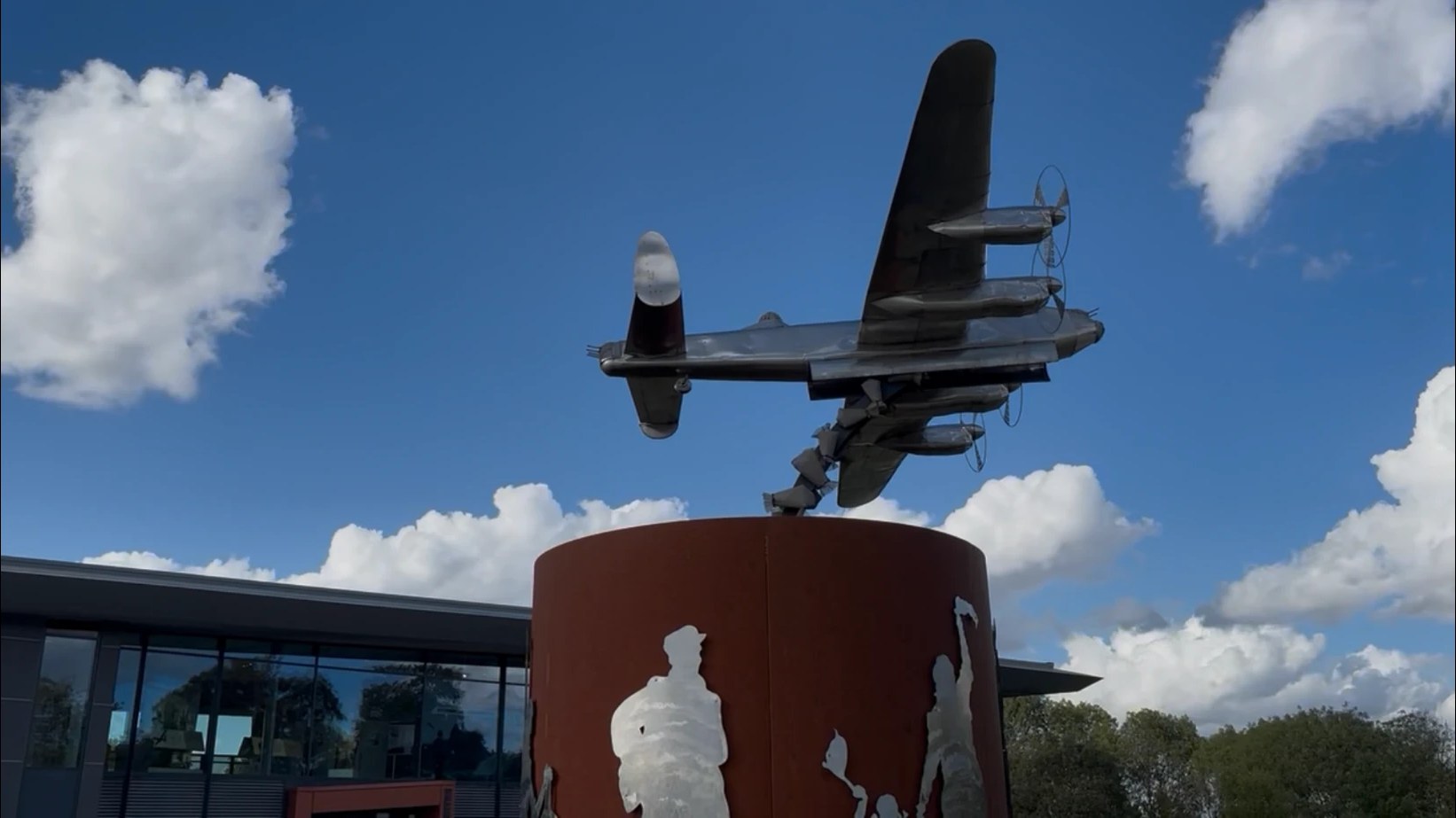 A stainless-steel Lancaster bomber sculpture on a rust-coloured Corten steel plinth with cut-out figures, outside the International Bomber Command Centre, against a blue sky with white clouds.