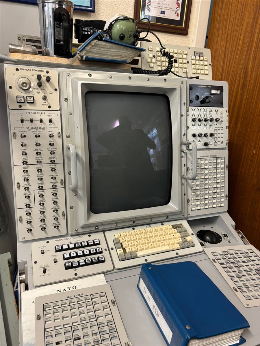 Grey 1970s-80s radar operator console with a dark CRT screen, switch panels, a keyboard and a blue binder.